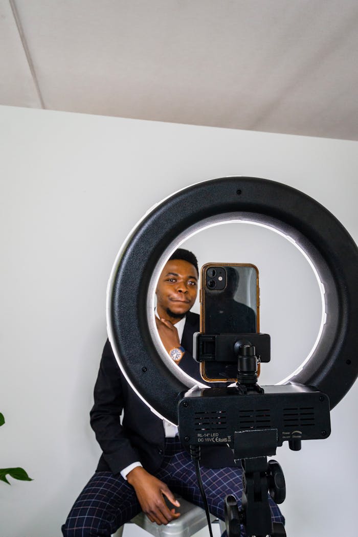 Man in black suit using a smartphone ring light for professional headshot, seated indoors.